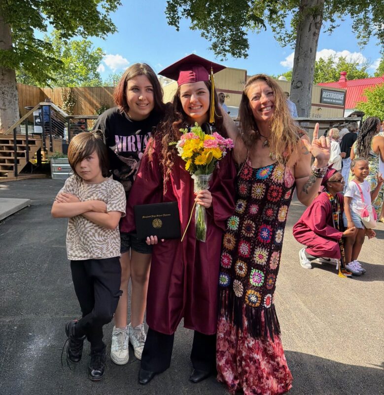 Aradia R. on graduation day in her cap and gown, holding a bouquet of flowers, surrounded by family and her mentor.