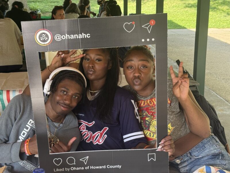 OhanaHC students smiling together under a pavilion at an outdoor community event, one holding up a photo frame prop.