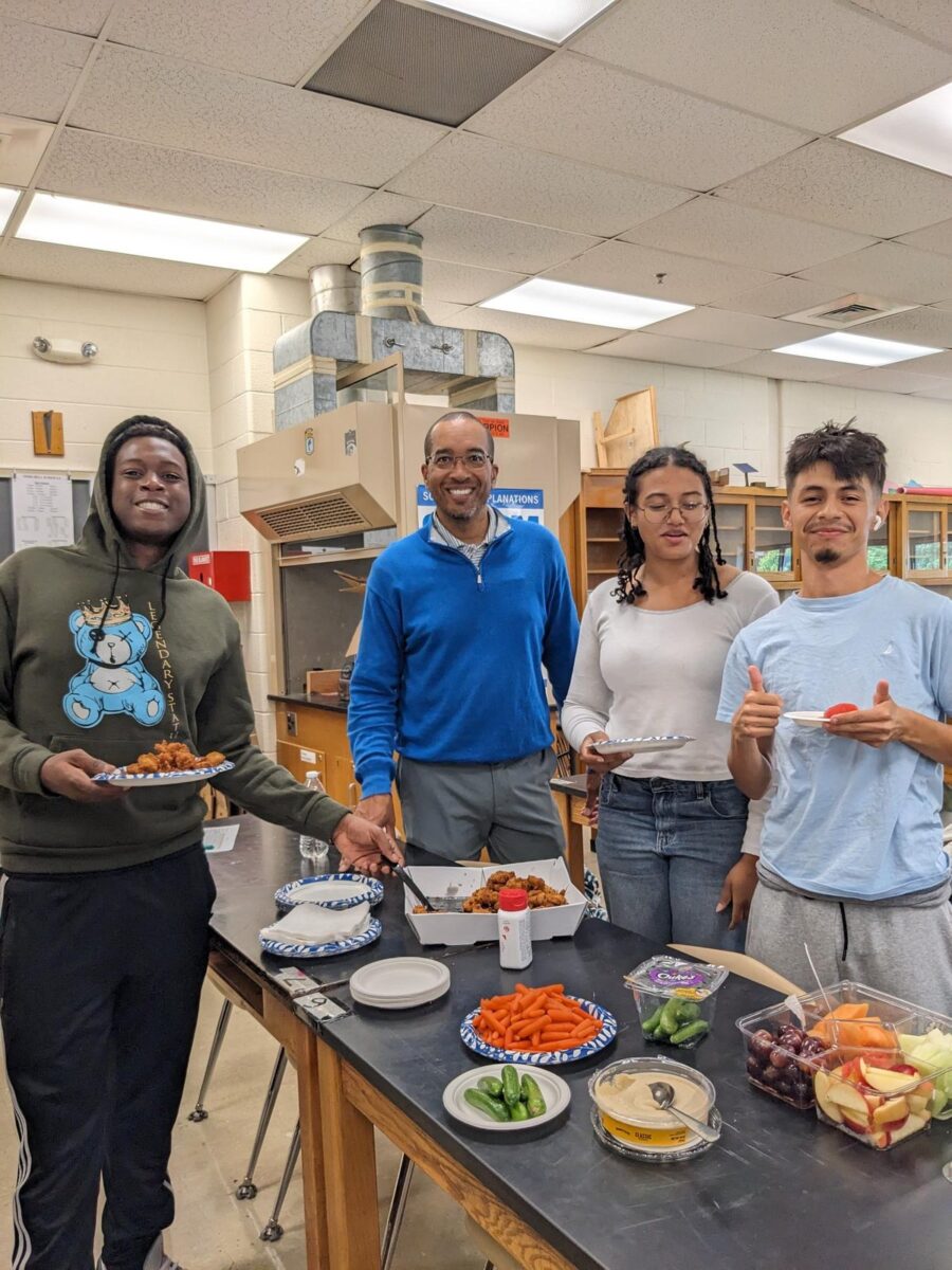 Three Oakland Mills students and an OhanaHC mentor smiling around a science lab table set with snacks at a club meeting
