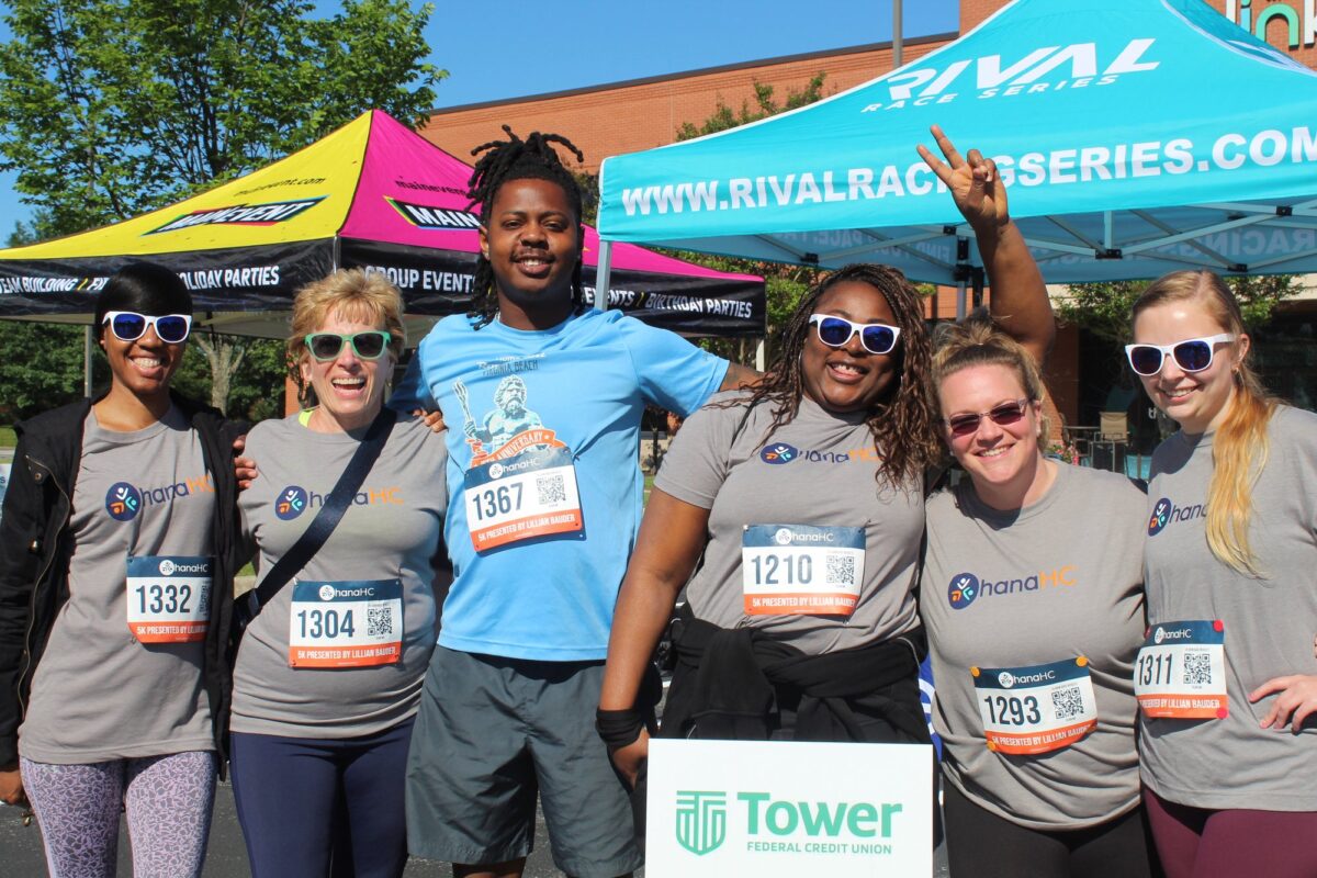 A team of runners in OhanaHC race shirts and bibs celebrating together at the Ohana 5K, beside a Tower Federal Credit Union sign.