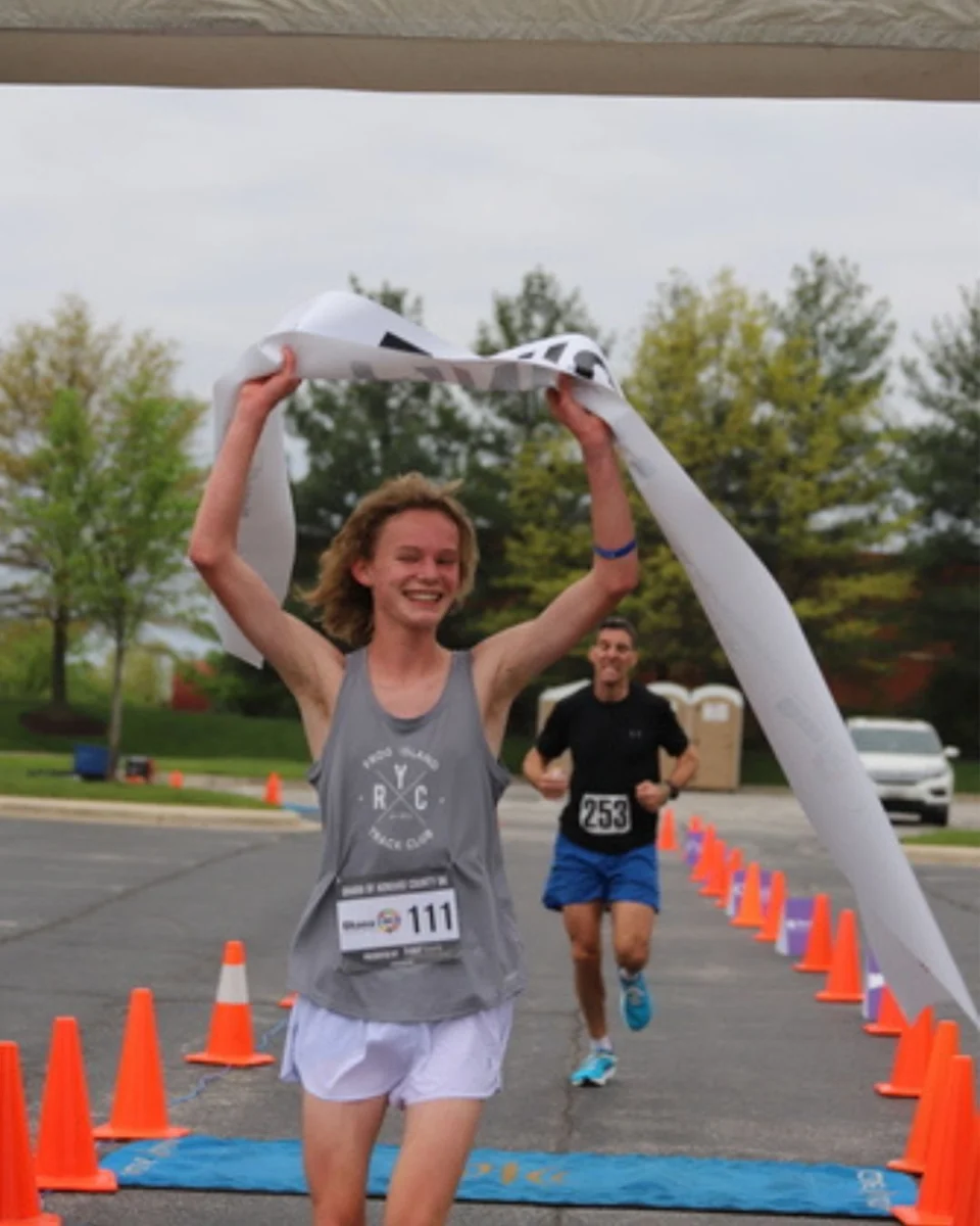 A runner breaking the tape at the Ohana 5K with her arms raised, the next runner coming in behind her.