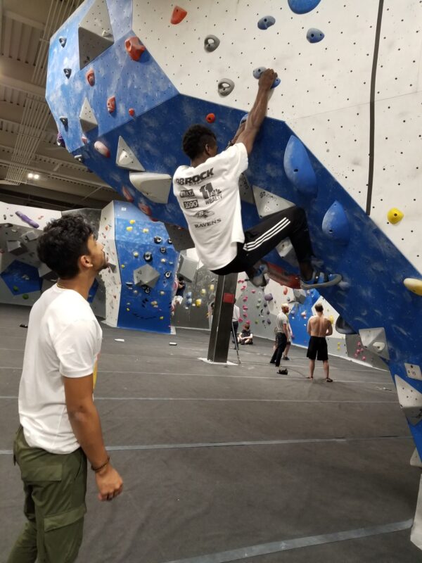 A young person part way up an indoor climbing wall while another watches from the floor