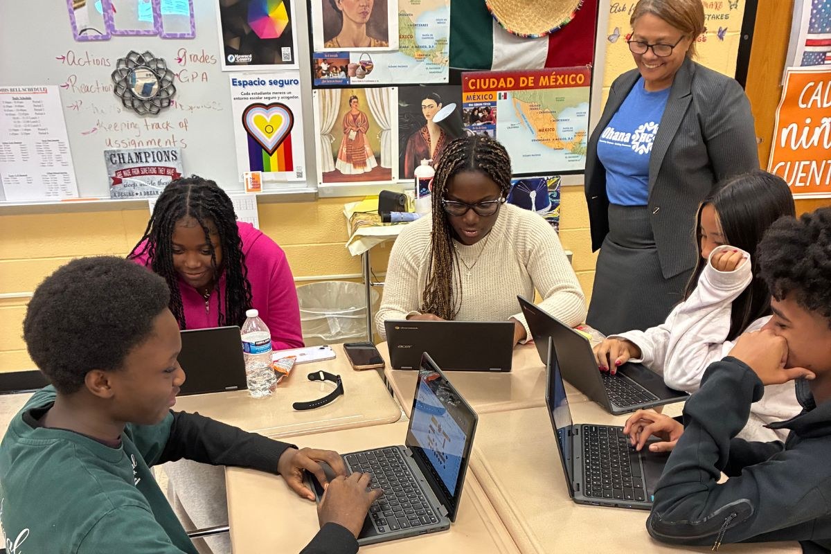 Oakland Mills students working on laptops around a classroom table while an OhanaHC staff member looks on and smiles